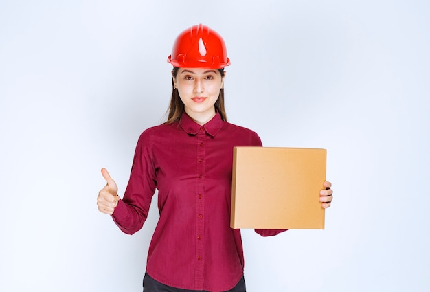 Фото женщины в красном шлеме держа картонную коробку и давая большие пальцы руки вверх.
