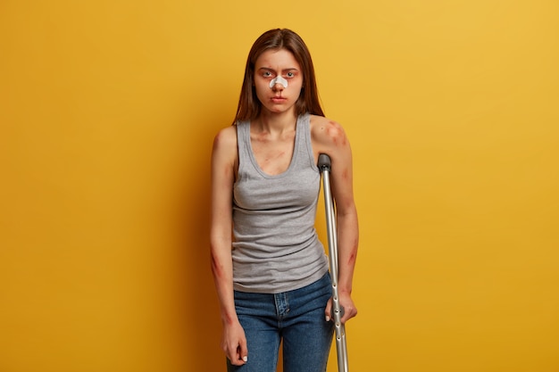 Free photo photo of woman victim of accident has nose breakage, poses with crutch, cannot walk by herself, gets concequences of careless driving, dressed in vest, jeans, has abrasion and bruises on skin