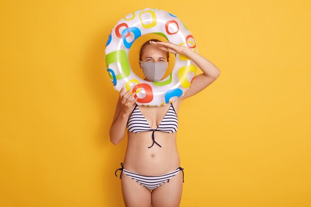 Photo of woman wearing protective mask posing isolated over rosy wall, lady with inflatable ring, keeping hand near forehead and looking in distance.