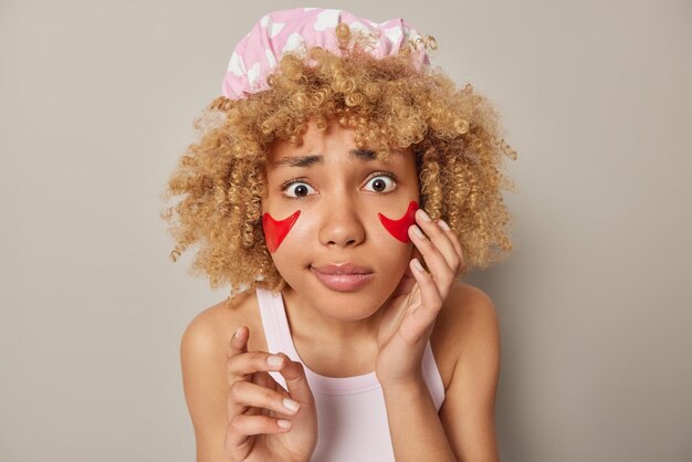 Фотография взволнованной кудрявой женщины в шляпе для ванны и повседневной футболке с красными пятнами под глазами, заботится о нежной коже, изолированной на сером фоне. Косметологические процедуры