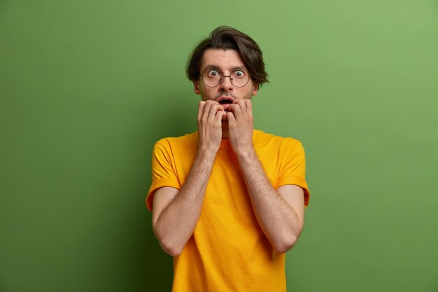 Фотография встревоженного нервного мужчины кусает ногти и смотрит с испуганным выражением лица, испуганный чем-то ужасающим, в круглых очках и желтой футболке, позирует у зеленой стены.