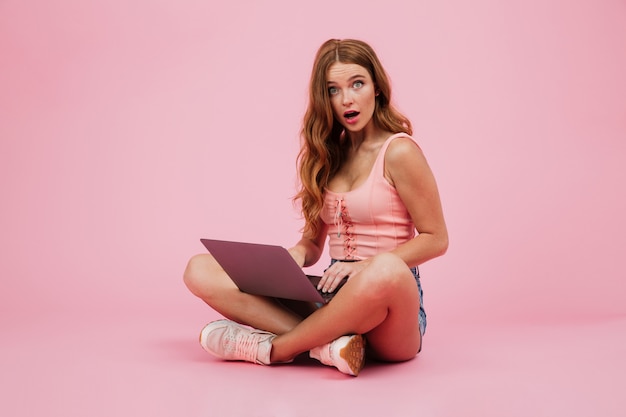 Photo of young amazed readhead woman with laptop, siiting with crossed legs