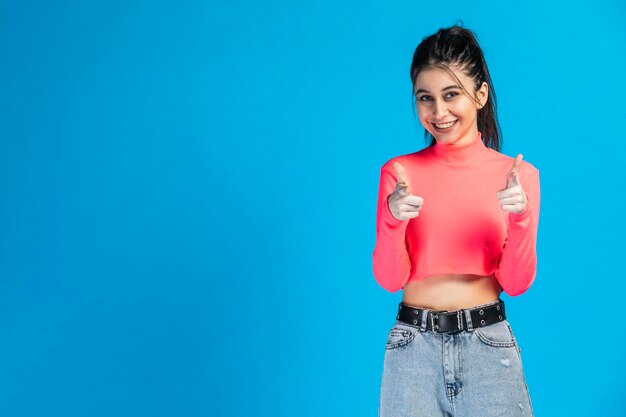Photo of young beautiful girl standing on blue background and point her fingers to the camera