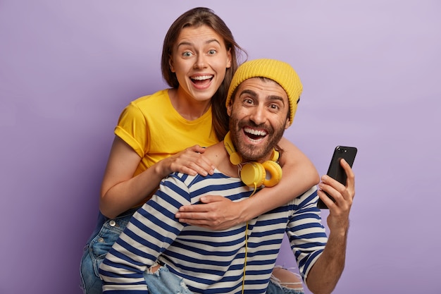 若いボーイフレンドとガールフレンドの写真は一緒に楽しんでいます、男性は女性にピギーバックの乗り物を与え、携帯電話を使用し、楽しく笑い、紫色の壁に隔離されています。幸せなブロガー
