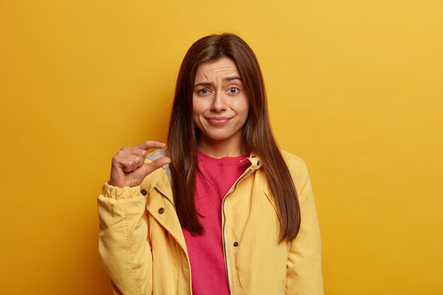 Photo of young dark haired woman shows very small size with fingers, demonstrates tiny measure, tells about decreased prices and little salary, raises eyebrows with bewilderment, wears yellow jacket