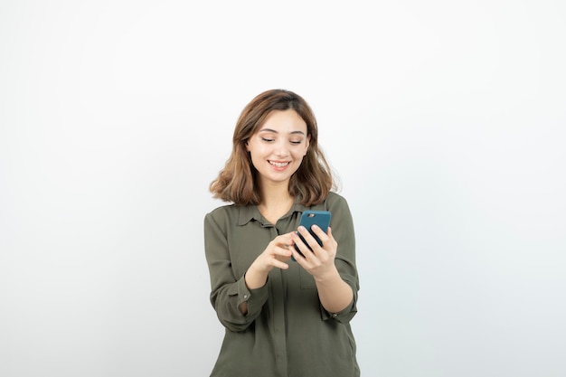 Фотография молодой девушки с мобильным телефоном, стоящей над белой стеной. Фото высокого качества
