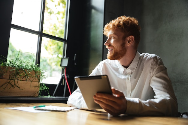 Photo of young handsome readhead bearded man holding tablet, looking at big window