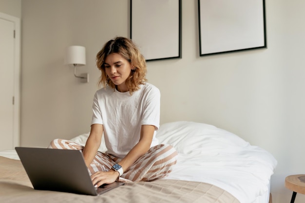 Free photo photo of young lady with wavy hairstyle wearing white pyjamas is sitting in the bed in the morning and working on laptop in sunlight in modern stylish apartment