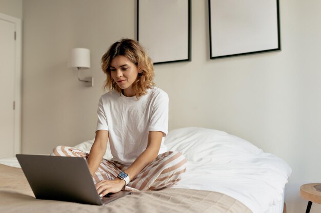 흰색 잠옷을 입은 물결 모양의 헤어스타일을 한 젊은 여성의 사진이 아침에 침대에 앉아 햇빛 아래 현대적인 세련된 아파트에서 노트북 작업을 하고 있습니다