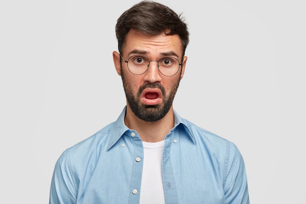 Photo of young male with dark stubble makes bored face, listens something with lack of interest, has discontent expression, dressed in blue shirt, opens mouth in displeasure, isolated over white wall