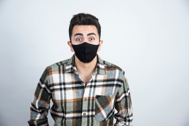 Фотография молодого человека в черной маске для защиты от коронавируса, стоящего над белой стеной.