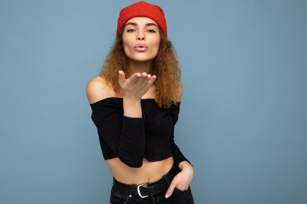 Photo of young positive cute nice brunette woman curly with sincere emotions wearing stylish black crop top and red hat isolated on blue background with copy space and giving kiss.