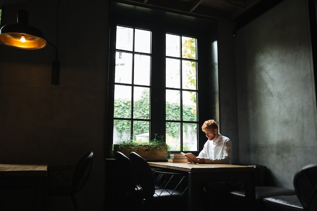 Photo of young redhead bearded man in white shirt reading a book in cafeteria