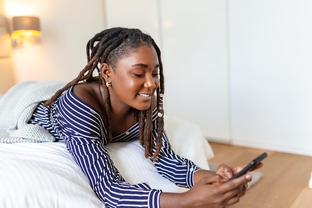 Photo of a young smiling African woman has a video call at night while lying on a bed in her bedroom