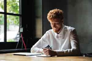 Free photo photo of young smiling readhead bearded man, taking notes, while sitting at cafe