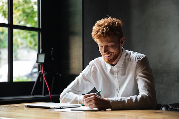 Photo of young smiling readhead bearded man, taking notes, while sitting at cafe