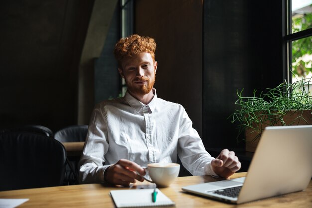 Photo of young smiling redhead bearded man holding coffee cup at cafeteria