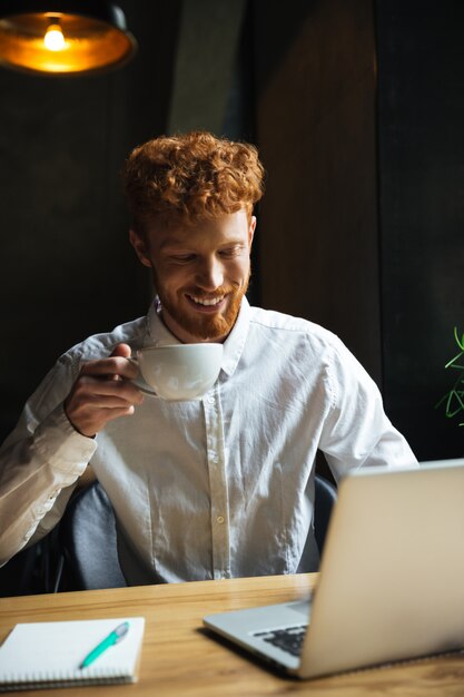 Foto gratuita foto della tazza di caffè sorridente della tenuta dell'uomo barbuto della giovane testarossa, esaminante lo schermo del computer portatile mentre lavorando al self-service