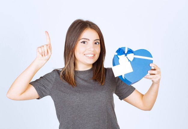 Photo of young smiling woman pointing a finger up and holding a heart shaped gift box.