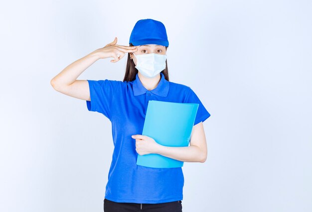 Photo of young woman in medical mask holding folder and pointing finger at her head. 