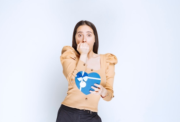 Photo of a young woman model holding a heart shaped gift box against white wall.