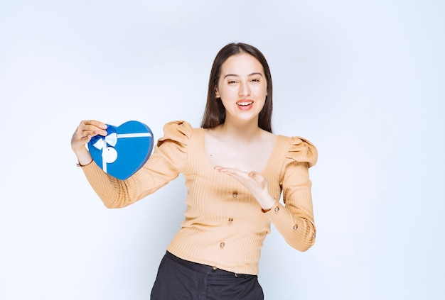 Photo of a young woman model pointing at a blue gift box against white wall.