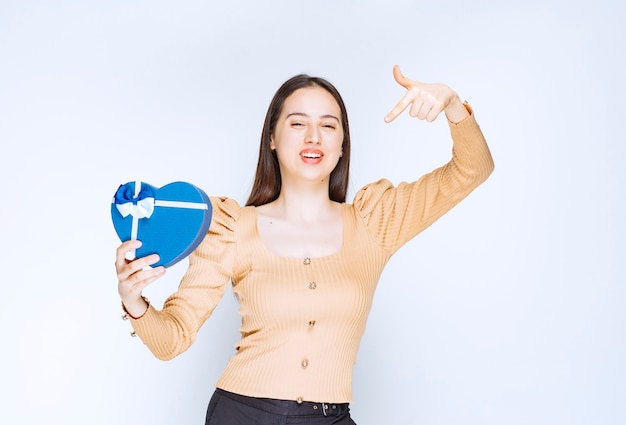 Photo of a young woman model pointing at a heart shaped gift box against white wall.