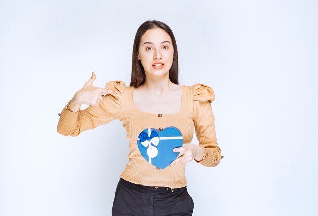 Photo of a young woman model pointing at a heart shaped gift box against white wall.