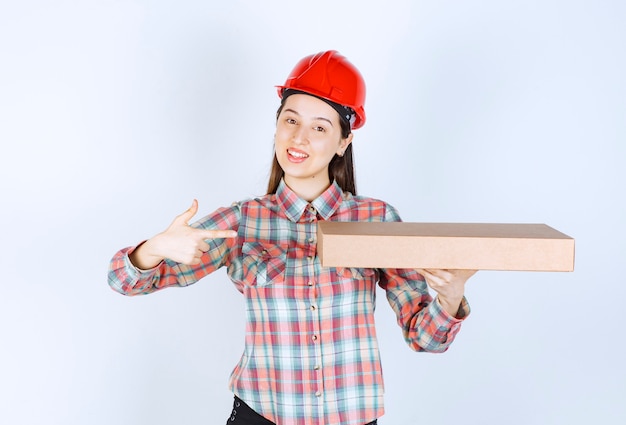 Photo of young woman in red helmet holding carton box and pointing. 