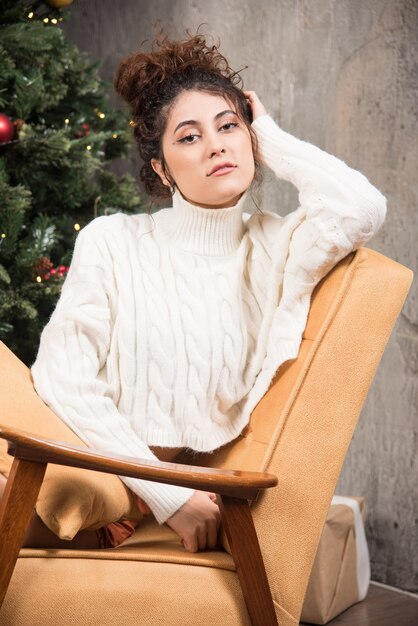クリスマスツリーの近くの快適な椅子に座っている若い女性の写真