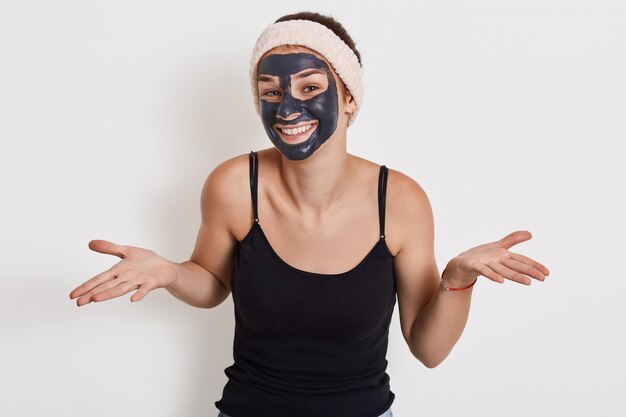Фотография молодой женщины с очаровательной улыбкой, разводящей ладони в стороны, показывает беспомощный жест, носит питательную лицевую маску для уменьшения морщин, позирует на белой стене.
