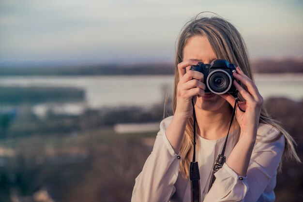Фотограф женщина девушка держит dslr фотокамера фотографирует. Улыбается молодая женщина, используя камеру, чтобы сделать фотографию на открытом воздухе