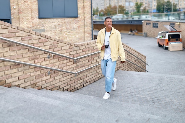 Photographer. Young dark-skinned smiling man with camera around your neck in light clothes walking on street stairs looking to side on fine day