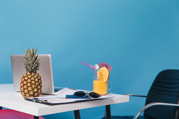 Foto del posto di lavoro durante le vacanze. ananas, cocktail arancione, bicchieri, grafica, laptop sono sul tavolo.