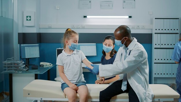 Physician consulting little girl with arm injury at exam appointment. General practitioner examining bones fracture of child in medical cabinet. Checkup visit during covid 19 pandemic.