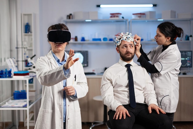 Physician doctor with virtual reality headset while neurologist woman adjusting eeg scanner of man patient analyzing brain evolution during neurology experiment. Scientist engineer using high tech