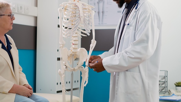 Free photo physician examining human skeleton in cabinet at checkup visit, explaining anatomy bones diagnosis to elderly patient. specialist analyzing spinal cord to help with orthopedic treatment.