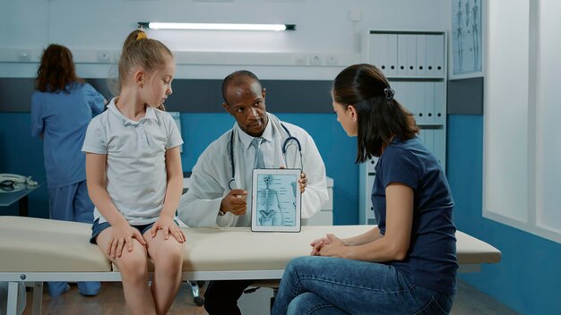 医師がデジタルタブレットで人間の骨格画像をオフィスの小さな女の子と親に説明します。骨の損傷と痛みを示すために小さな子供とオステオパシーの相談をしている医師。