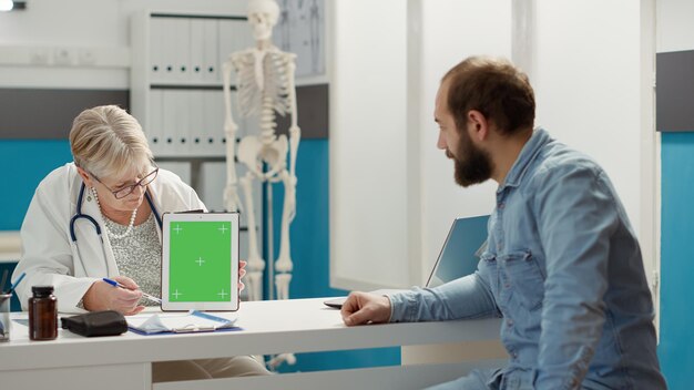 医師と患者は、診療所でグリーンスクリーンを備えたデジタルタブレットを分析しています。孤立したクロマキーの背景とテンプレートを持つ空白のコピースペースのモックアップを使用して男と薬。三脚ショット。