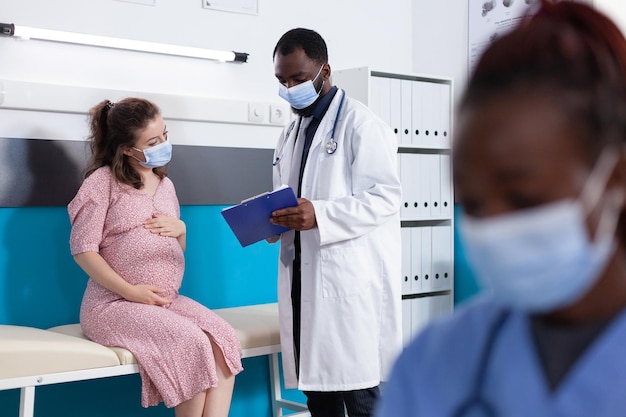 Physician showing checkup papers to pregnant woman in medical office. General practitioner doing examination with patient expecting child to check healthcare while wearing face masks