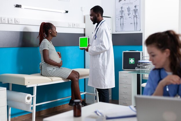 妊娠中の女性に垂直緑色の画面でタブレットを示す医師。大人は、ヘルスケアのための分離されたテンプレートとモックアップの背景を持つクロマキーを見ている赤ちゃんを期待しています。