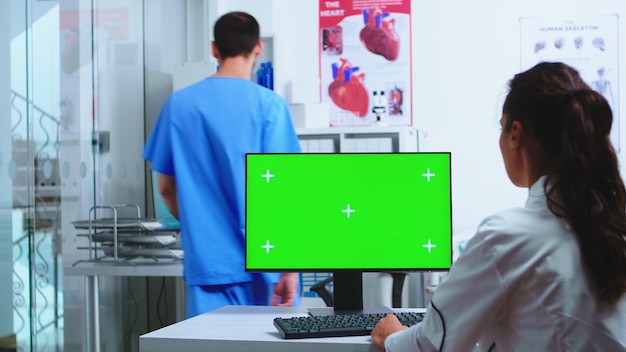 Physician using computer with chroma key and assistant holding patient x-ray in hospital cabinet. Desktop with replaceable green screen in medical clinic while doctor is checking patient radiography f