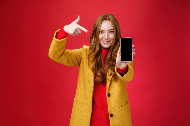 절대 후회하지 않는 휴대폰을 선택하세요. 노란색 코트를 입은 친근하고 매력적이며 자신감 넘치는 빨간 머리 매력적인 여자 친구의 초상화는 스마트폰이 카메라를 향해 활짝 웃는 것처럼 모바일을 가리키고 있습니다.