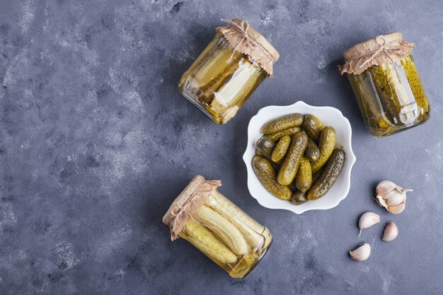 Pickled cucumbers in bowl and glass jars on blue background with garlic.