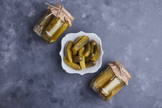 Pickled cucumbers in bowl and glass jars on blue background.