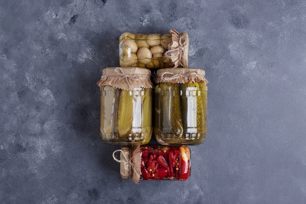 Pickled cucumbers, olives and red peppers in glass jars on blue background.