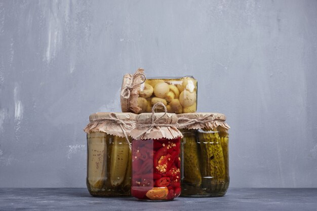 Pickled cucumbers, olives and red peppers in glass jars on blue background.