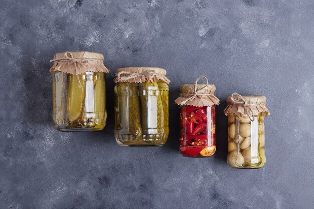 Pickled cucumbers, olives and red peppers in glass jars on blue background.