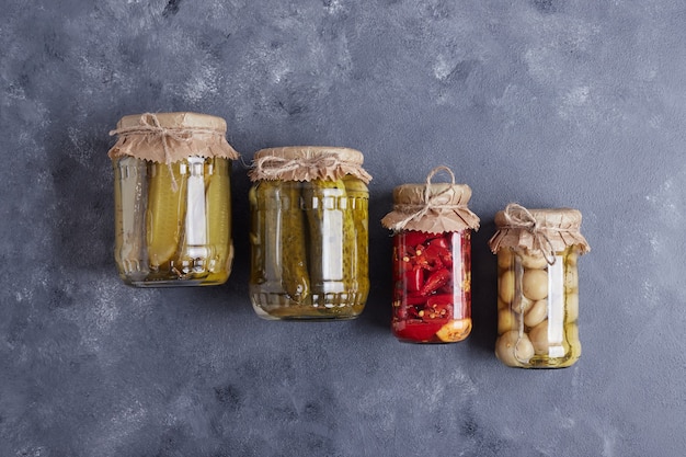 Pickled cucumbers, olives and red peppers in glass jars on blue background.