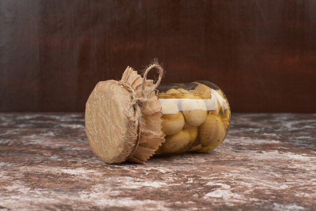 Pickled mushrooms in a glass jar on marble table. 